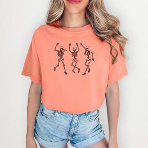 May include: Coral-colored t-shirt featuring a black graphic of three dancing skeletons. The skeletons are depicted in a playful pose, with their arms raised and legs in motion. The shirt is made of a soft material and has a relaxed fit.