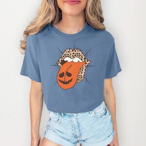 May include: A blue t-shirt featuring a Halloween-themed graphic. The graphic depicts a mouth with leopard print lips and a tongue shaped like a jack-o'-lantern. A spider web design surrounds the mouth.