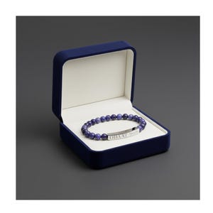 May include: A blue velvet jewelry box with a white interior holds a beaded bracelet. The bracelet features dark blue beads and a silver bar engraved with the word "ODAAT". The box is open, displaying the bracelet.