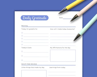 May include: A white daily gratitude planner sheet with blue text and lines, featuring sections for morning goals, affirmations, and a night-time review. Three pens in yellow, light pink, and light mint green are positioned at the top right corner.