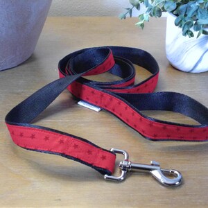 May include: A red and black dog leash with a star pattern. The leash has a silver metal clasp and is lying on a wooden surface. A white tag is attached to the leash. A decorative plant is in the background.