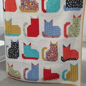 May include: A rectangular quilt featuring a grid of sixteen cat-shaped fabric appliques in various colors and patterns, including red, blue, yellow, and floral prints, set against a white background with a yellow border and visible grid stitching.