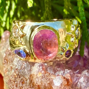 Könnte beinhalten: Ein goldener Ring mit einem großen, ovalen, rosa Edelstein im Zentrum. Der Ring ist mit kleineren blauen und violetten Edelsteinen sowie dekorativen goldenen Details verziert. Der Ring ist vor grünem Laub und einer kristallinen Oberfläche platziert.