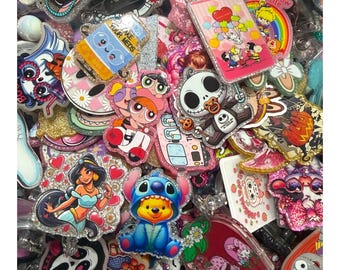 Puede incluir: Una colección de coloridos y brillantes carretes para insignias con varios personajes y diseños de dibujos animados. El surtido incluye imágenes de Jasmine, Stitch, Jack Skellington y las Supernenas, entre otros. Los carretes para insignias tienen una variedad de formas y tamaños.
