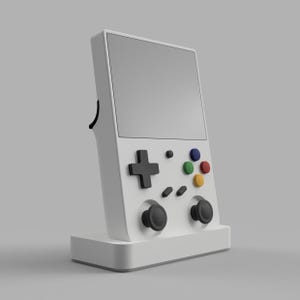 May include: A white handheld gaming console with a black screen, a black directional pad, and four colored buttons. The console is on a white stand.