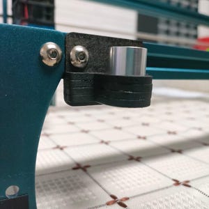 May include: Close-up of a teal blue 3D printer with a black and silver metal bracket attached to the side. The bracket has two silver screws and a silver metal tube.