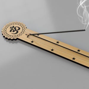 Puede incluir: Un porta incienso de madera clara con un símbolo Om decorativo y recortes de estrellas. El soporte tiene bordes negros y está diseñado para sostener un palito de incienso encendido. El humo blanco se eleva del incienso.