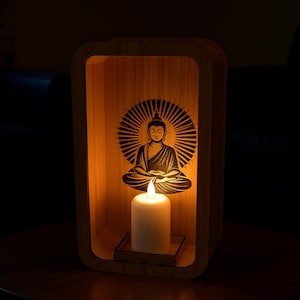 May include: A wooden Buddha candle holder with a glowing candle. The holder has a rectangular shape with a Buddha silhouette on the back. The candle is white and sits on a small wooden tray. The overall color scheme is warm and inviting.