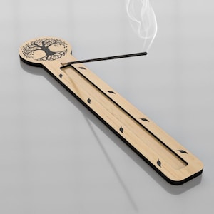 Può includere: Un porta incenso in legno con un design ad albero della vita. Il supporto è rettangolare, con finitura in legno chiaro e bordi neri. Un bastoncino di incenso è acceso, con fumo che sale. Il design include ritagli a forma di foglia sui lati.