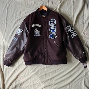 Peut inclure: Veste universitaire Champion marron avec des manches en cuir marron. La veste présente le mot "Champion" et l'année "1919" en lettres blanches. Des écussons et les lettres "CCC" sont également visibles.