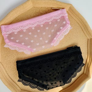 May include: Two pencil cases shaped like underwear. One is pink with white heart patterns and lace trim. The other is black with heart patterns and lace trim. Both have a zipper closure.