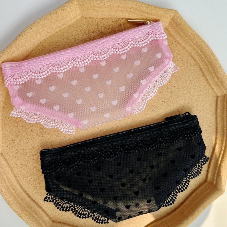 May include: Two pencil cases shaped like underwear. One is pink with white heart patterns and lace trim. The other is black with heart patterns and lace trim. Both have a zipper closure.