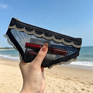 May include: Black mesh cosmetic bag with gold trim and a gold zipper. The bag is filled with lipsticks and other makeup items. The bag is held up against a beach background.