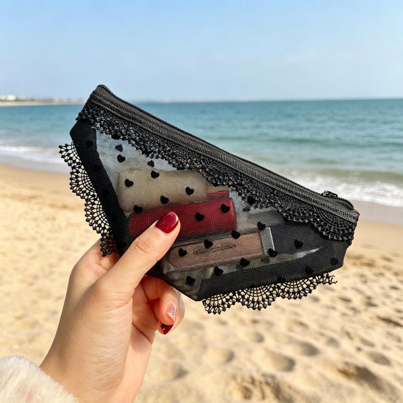 May include: A black mesh cosmetic bag with a zipper closure, adorned with small heart patterns and lace trim. The bag contains several lipsticks in various shades. The background shows a beach with the ocean.