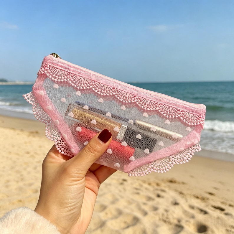 May include: Pink lace cosmetic bag with white polka dots, holding lipsticks and makeup tools. The bag has a gold zipper and a delicate lace trim. The bag is made of a sheer material, allowing the contents to be partially visible.