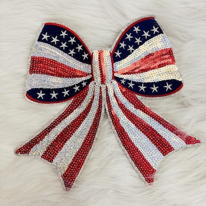 May include: A large, decorative bow featuring a patriotic design. The bow is adorned with red, white, and blue sequins, mimicking the American flag. The design includes red and white stripes, along with a blue section with white stars. The bow is likely for festive occasions.
