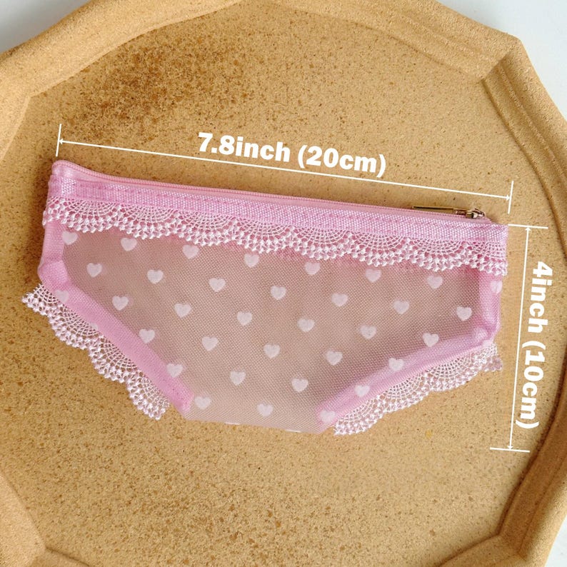 May include: A pink, heart-patterned pouch, 7.8 inches (20 cm) long and 4 inches (10 cm) high, with a lace trim. The pouch has a zipper closure and is made of a sheer material. The pouch is on a light brown surface.