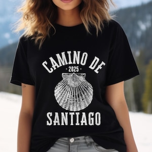 May include: Black t-shirt with the text "CAMINO DE SANTIAGO - 2025" in white, featuring a detailed seashell graphic. The design is centered on the shirt, with the words arched above and below the shell.
