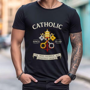 May include: Black t-shirt with the word "CATHOLIC" above a crest featuring keys, a crown, and the text "SINCE 33 AD." A banner below reads "ONE, HOLY, CATHOLIC, AND APOSTOLIC CHURCH."