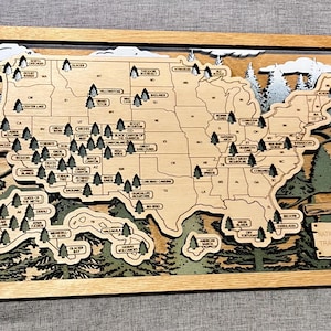 Peut inclure: Une carte en bois encadrée des États-Unis avec des découpes superposées des frontières des États et des parcs nationaux. De petites icônes d'arbres marquent les parcs sur la carte, incluant l'Alaska et Hawaï. Un panneau indique National Park Travel Guide.