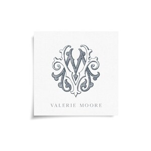 May include: A square card with a detailed, gray-blue monogram design featuring the letters "V" and "M" intertwined within an ornate, floral-inspired frame. The name "VALERIE MOORE" is printed in a simple font below the design.