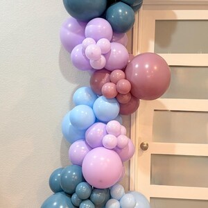 May include: A balloon arch in shades of blue, lavender, and mauve, arranged in a cascading design. The balloons vary in size, creating a textured and visually appealing display. Perfect for a party or event.