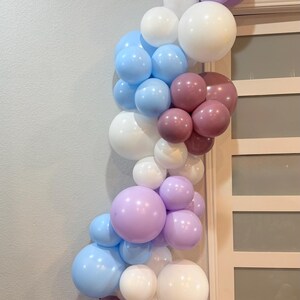 May include: A balloon garland in shades of lavender, light blue, white, and mauve. The balloons are arranged in a cascading, organic shape, perfect for a party or event decoration. The garland is against a neutral wall and door.