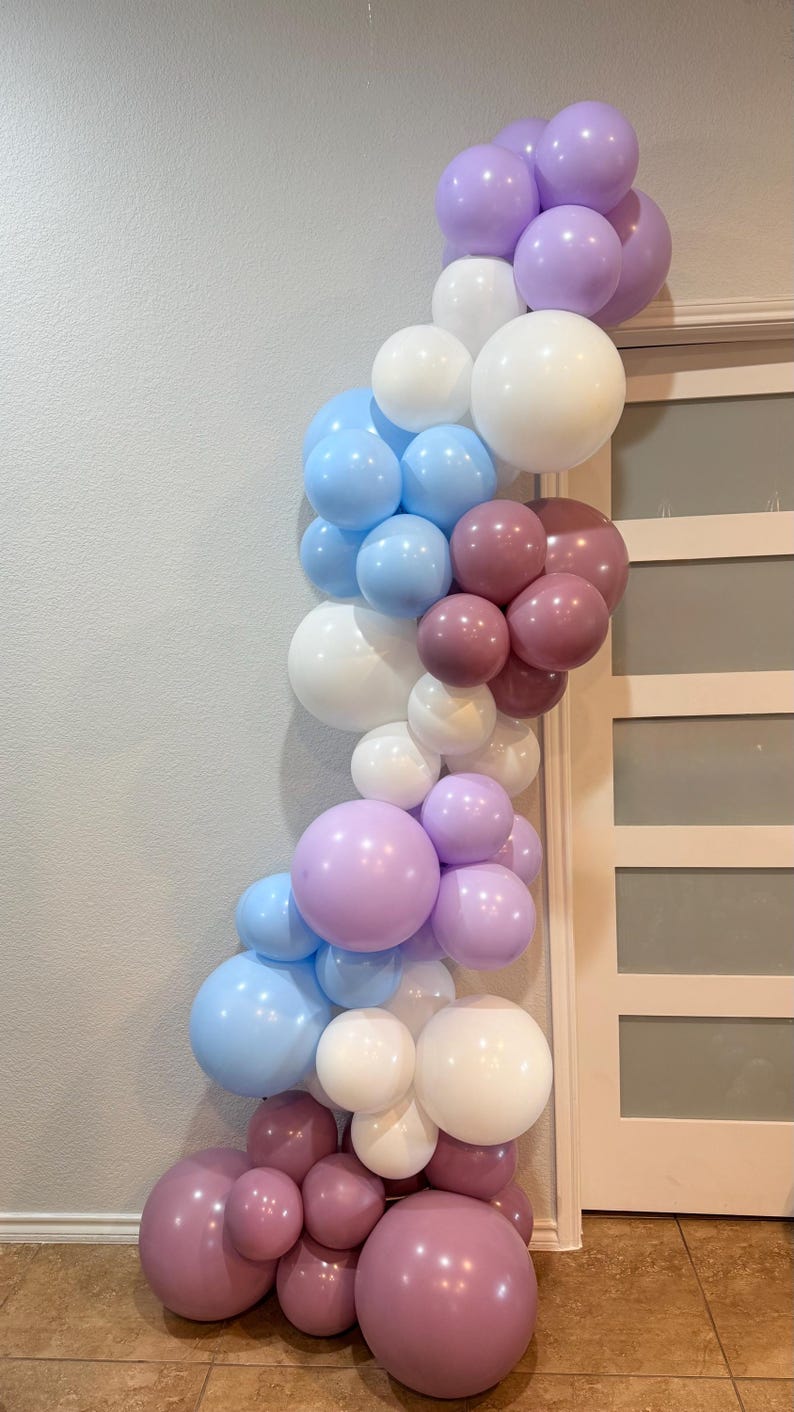 May include: A balloon garland in shades of lavender, light blue, white, and mauve. The balloons are arranged in a cascading, organic shape, perfect for a party or event decoration. The garland is against a neutral wall and door.