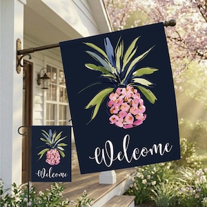 May include: Two navy blue outdoor flags with a watercolor pineapple design. The pineapple is pink and green, with the word "Welcome" in white script. The flags are hanging outside a house.