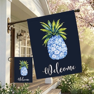 May include: A navy blue garden flag with a watercolor pineapple design. The pineapple is blue and white with green and yellow leaves. The word "Welcome" is written in white script. A smaller matching flag is also visible.