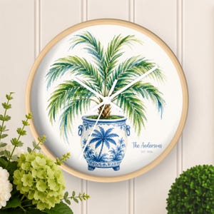 May include: A round wall clock with a light wood frame featuring a watercolor illustration of a green palm plant in a blue and white chinoiserie-style planter. The text The Andersons EST. 2026 is printed in blue script on the white clock face.