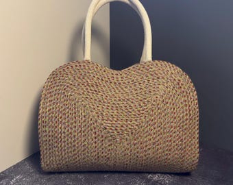 May include: A woven straw handbag with a white handle. The bag has a rounded, heart-shaped design and is decorated with a multi-colored pattern.