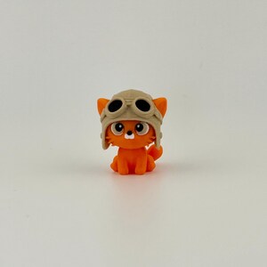 May include: An orange kitten figurine wearing a tan aviator helmet with black goggles. The kitten is sitting upright with its tail curled. The figurine has large, expressive eyes and a white patch on its nose. The background is plain white.