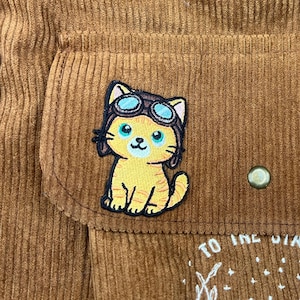 May include: Close-up of a brown corduroy fabric with a patch of a yellow cartoon cat wearing a pilot's helmet and goggles. The cat has blue eyes and is smiling. A button is visible on the right side of the image.