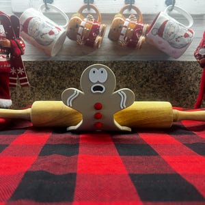 May include: A Christmas-themed image with a surprised gingerbread man, two wooden rolling pins, and festive mugs. The scene is set on a red and black plaid tablecloth, with Santa Claus figurines in the background. The mugs have Santa Claus designs.