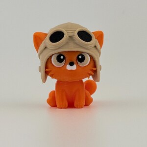 May include: An orange cat figurine wearing a tan aviator helmet with black goggles. The cat has large, round eyes and a white muzzle. The figurine is sitting on a white surface.