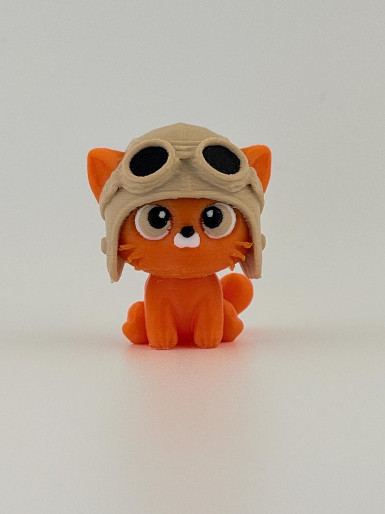 May include: An orange cat figurine wearing a tan aviator helmet with black goggles. The cat has large, round eyes and a white muzzle. The figurine is sitting on a white surface.