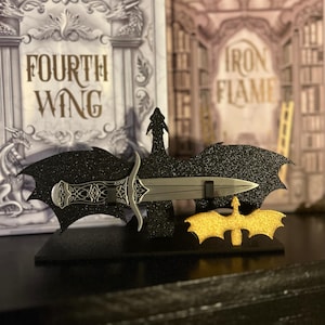 May include: A black glittery dagger display stand with a silver blade and decorative handle. The stand has a bat-wing design and a gold bat silhouette. The words "FOURTH WING" and "IRON FLAME" are visible in the background.