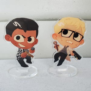 May include: Two acrylic character standees featuring stylized cartoon figures. The left figure has dark hair, a black and white plaid shirt, and holds a gold handgun. The right figure has blonde hair, black glasses, a white coat, and holds a black handgun.