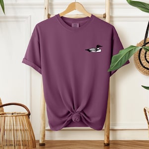 May include: A purple short-sleeved t-shirt with a black and white loon graphic on the front.