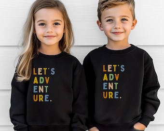 May include: Two children wearing black sweatshirts with the text "LET'S ADVENTURE" in colorful letters. The sweatshirts have a crew neck and long sleeves. The children are smiling and looking at the camera.