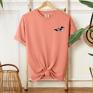 May include: A coral coloured t-shirt with a black and white loon graphic on the front.