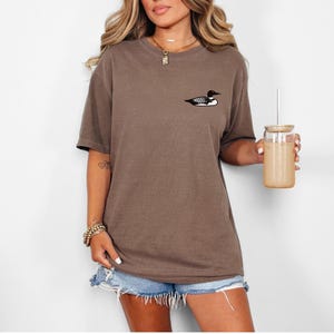 May include: An oversized brown t-shirt with a small black and white loon graphic on the front. The model is wearing the shirt with denim shorts and holding a glass with a straw.