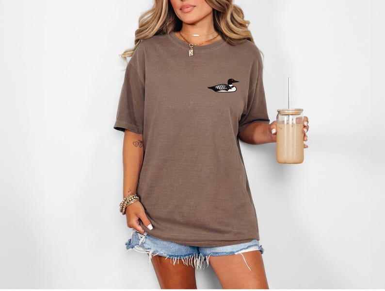 May include: An oversized brown t-shirt with a small black and white loon graphic on the front. The model is wearing the shirt with denim shorts and holding a glass with a straw.