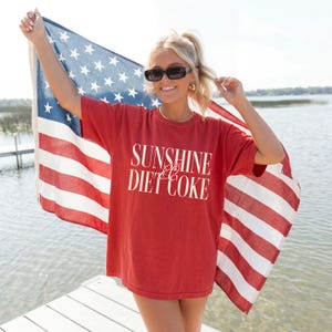 May include: Red t-shirt with the text "SUNSHINE & DIET COKE" in white. The person is holding an American flag. The background shows a body of water and a dock.
