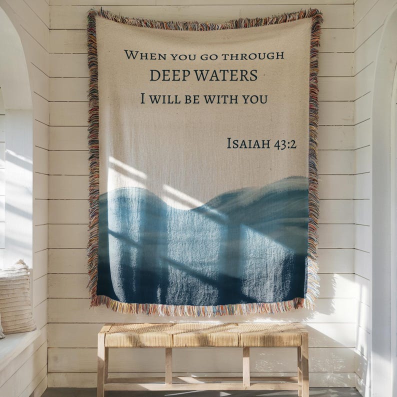 K&ouml;nnte beinhalten: Eine gewebte Wohndecke mit einem cremefarbenen oberen Bereich, der den schwarzen Text "WHEN YOU GO THROUGH DEEP WATERS I WILL BE WITH YOU ISAIAH 43:2" tr&auml;gt, und einem blauen Wellenmuster im Farbverlauf unten, mit mehrfarbigen Fransen am Rand.