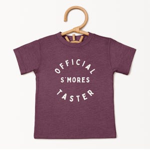 May include: A burgundy t-shirt with short sleeves, featuring the text "OFFICIAL S'MORES TASTER" in a white, curved font. The shirt is suspended on a light brown wooden hanger. The background is plain white. The fabric appears soft.