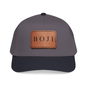 Puede incluir: Gorra de béisbol gris con visera azul marino. Un parche de cuero marrón con la palabra "BOJI" en letras negras está centrado en la parte delantera. La gorra tiene una visera curva y un botón en la parte superior.
