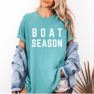 May include: A turquoise t-shirt with the words "BOAT SEASON" in bold white letters. The shirt is worn with a denim jacket and shorts. The overall style is casual and summery.