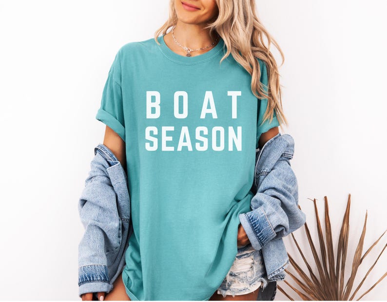 May include: A turquoise t-shirt with the words "BOAT SEASON" in bold white letters. The shirt is worn with a denim jacket and shorts. The overall style is casual and summery.